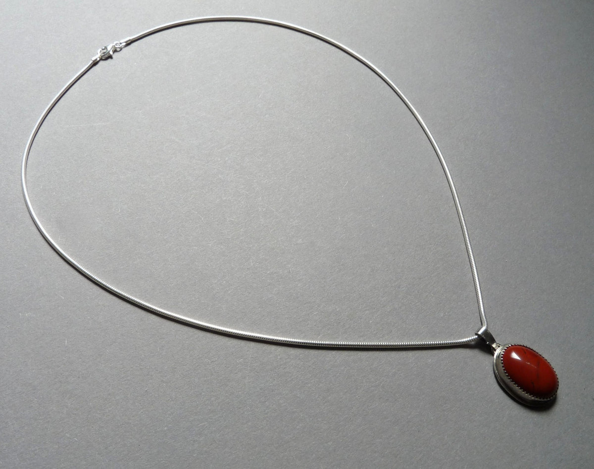 Handgemaakte zilveren slangenketting met een ovale rode Jaspis edelsteen hanger platliggend gefotografeerd op een grijze achtergrond