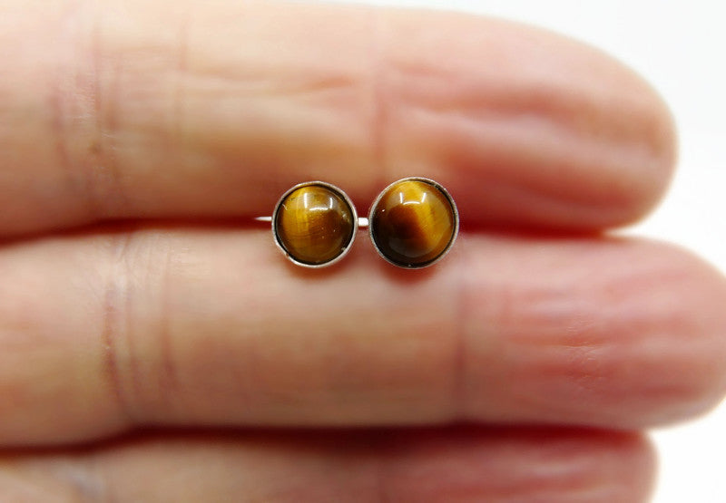 Twee bruine tijgeroog oorknopjes voor mannen vastgehouden in een hand om het 6mm formaat te tonen