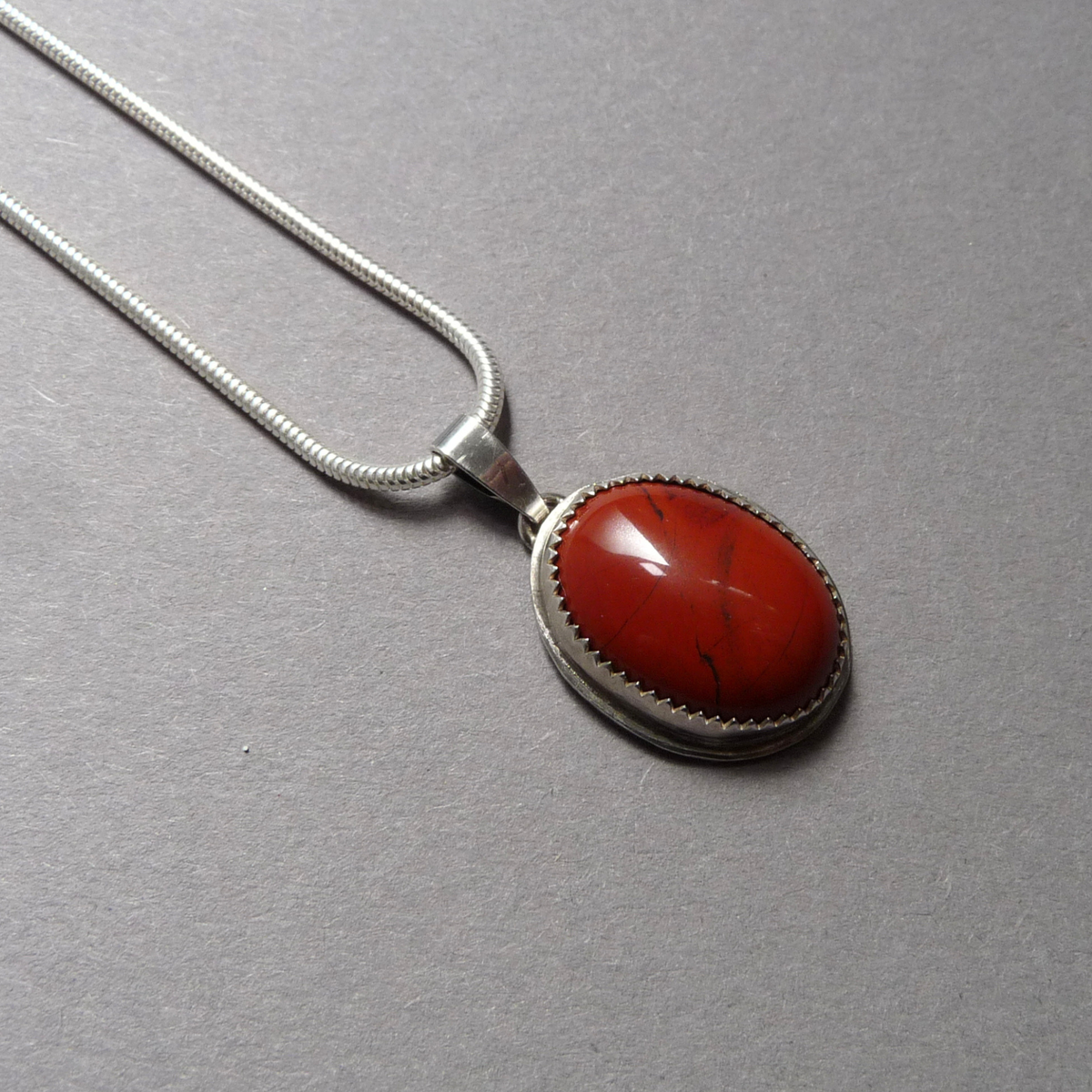 Zilveren damesketting met een glanzende rode Jaspis hanger liggend op een grijze ondergrond met focus op de edelsteen